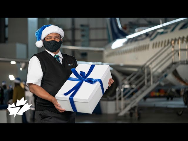 WestJetter wearing a blue Santa hat and a black mask, holding a gift in front of a WestJet plane